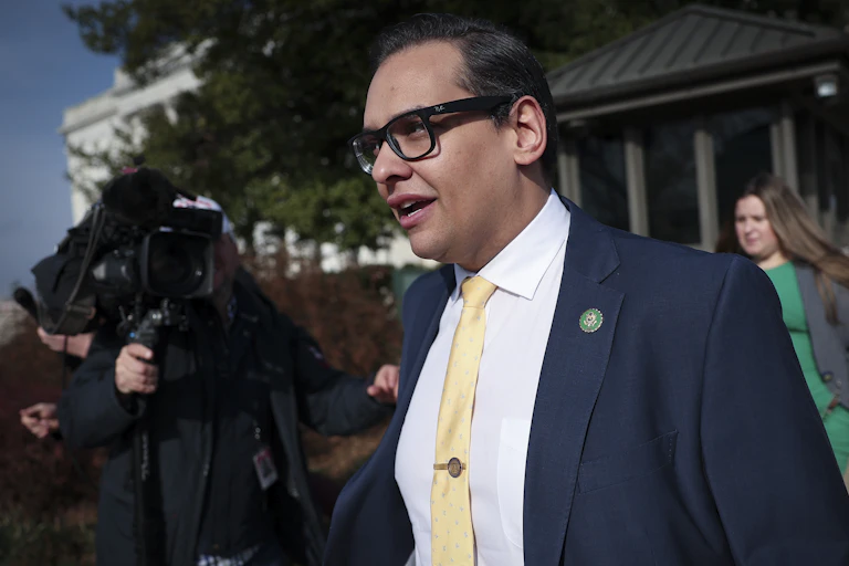 Man in a blue suit and yellow tie walks while being filmed by a large camera.