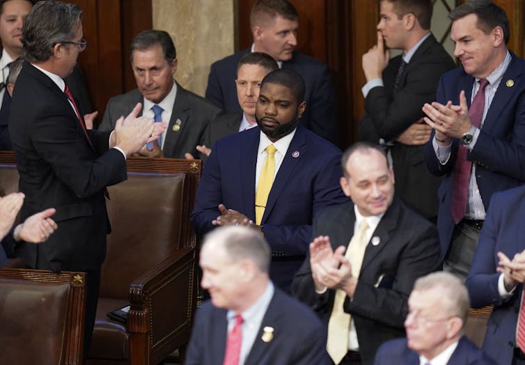 A black man dressed in a dark business suit is surrounded by several white men who are applauding