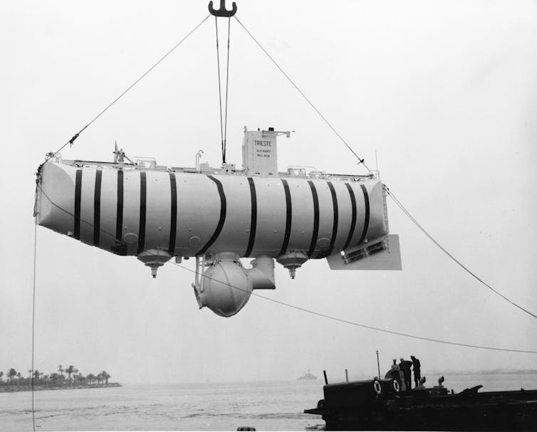 A black and white photo of a submarine with a round protrusion on the bottom, hanging in the air from a hoist.