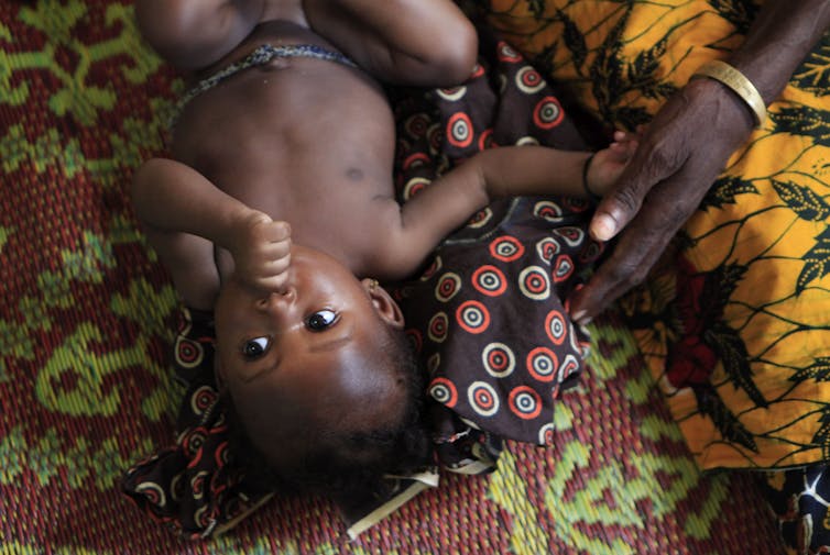 A baby sucking its thumb lies on its back on a patterned blanket.