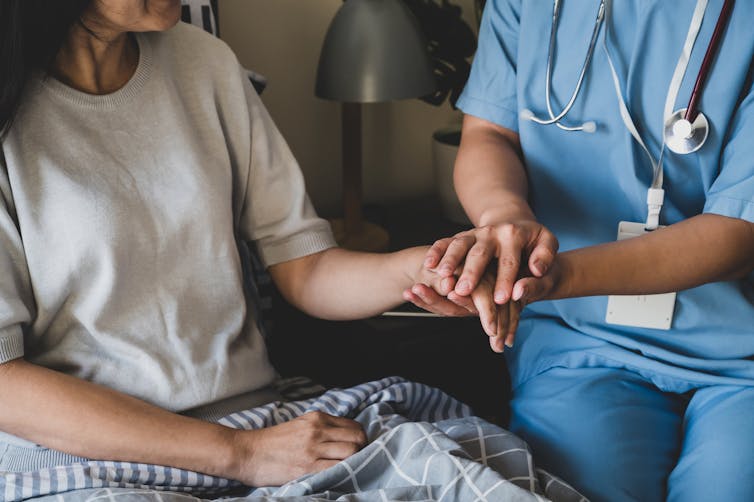 A woman in a white shirt sits in bed beside a doctor wearing a stethoscope who holds her hand.