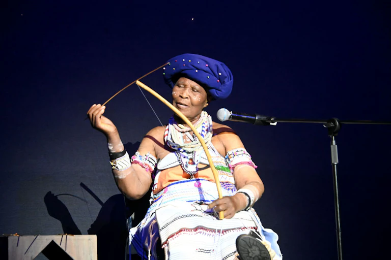 An elderly woman in traditional attire sits at a microphone. She holds a bow with a string in her lap and a thin stick in her hand.