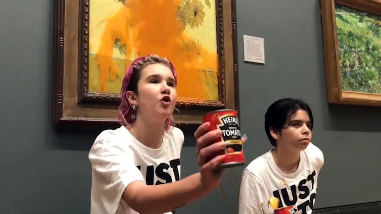 A protestor holds up an empty soup can in front of Van Gogh's sunflowers, which is covered in tomato soup.