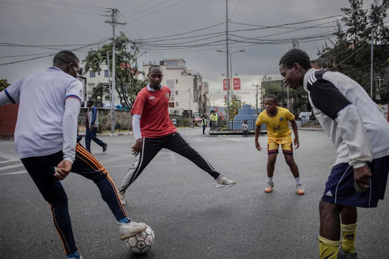 Four young men standing around each other, one about to kick a ball and the other three watching it closely.
