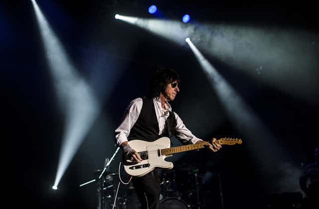 Jeff Beck wears a white shirt, black waistcoat and aviator sunglasses. He strums a white guitar.