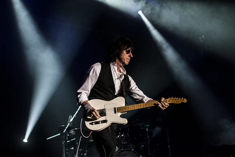 Jeff Beck wears a white shirt, black waistcoat and aviator sunglasses. He strums a white guitar.