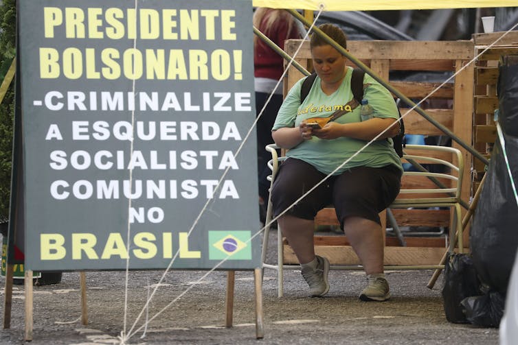 A woman sits looking at her phone outside a gate with a big pro-Bolsonaro banner.