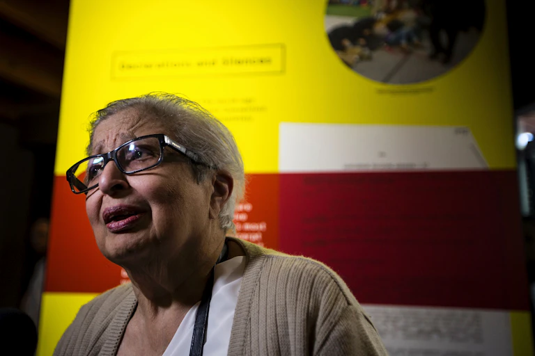 An elderly Indian woman with glasses and grey hair looks earnest as she speaks.