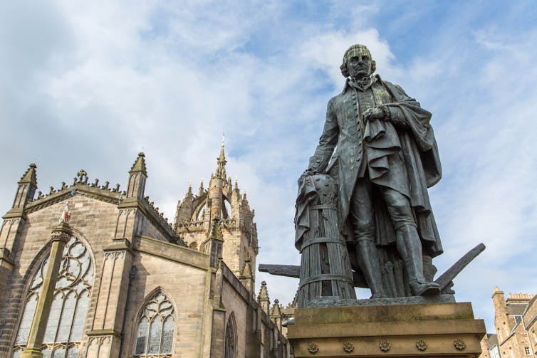 A statue of a man in Victorian clothing and a powdered wig standing on a pedestal in front an ornate building