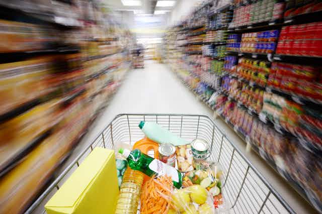 A point-of-view photograph of a shopping cart, speeding down a supermarket aisle, showing shelves on both sides filled with packaged foods.