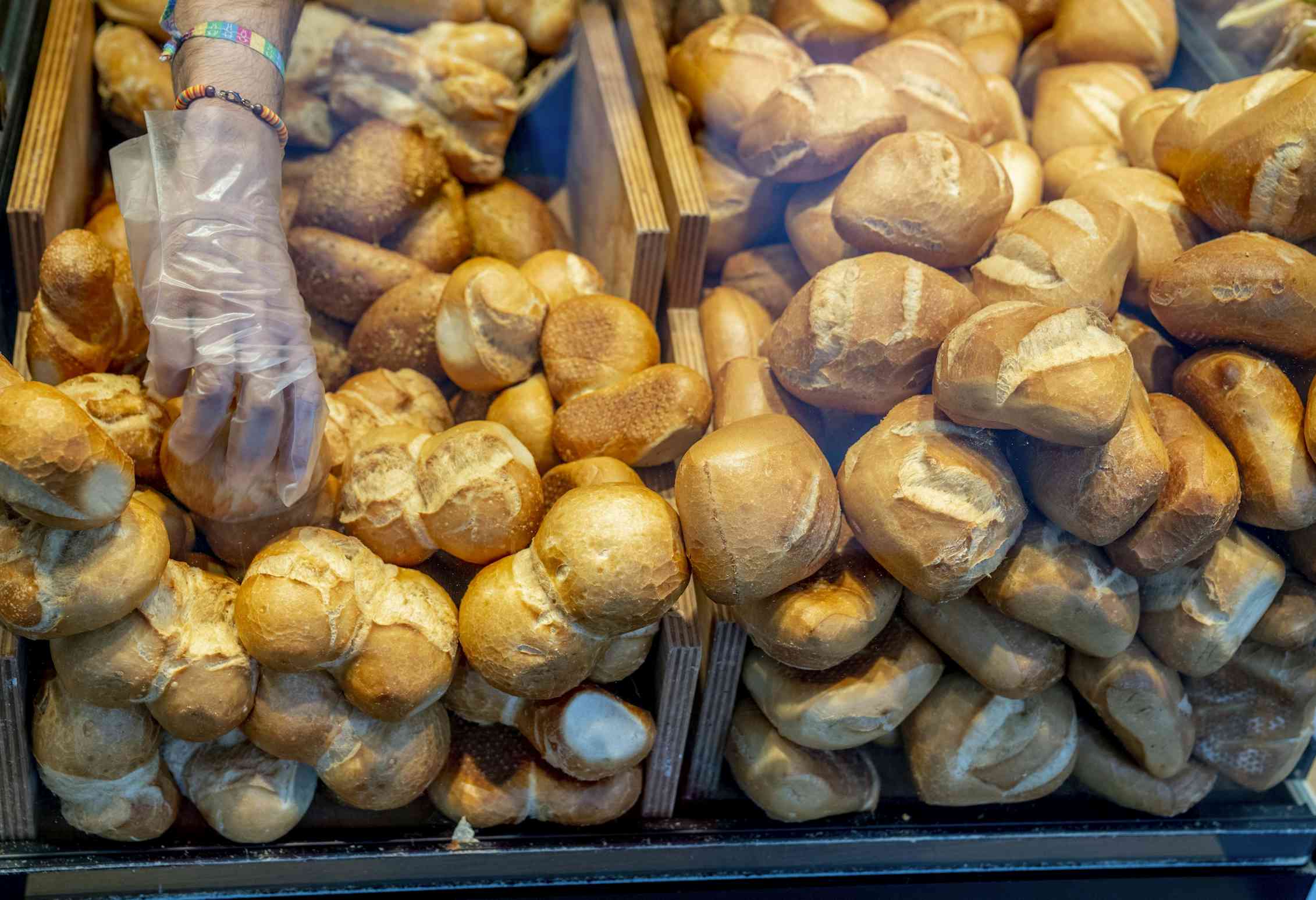 A gloved hand reaches into a bakery display case to pick up a bread bun from a pile