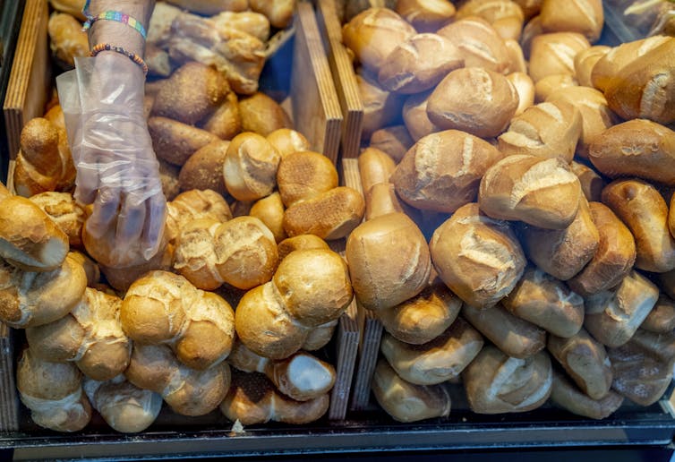 A gloved hand reaches into a bakery display case to pick up a bread bun from a pile