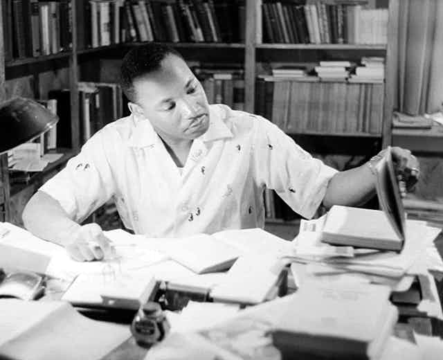 A black man sits at desk filled with papers and books.