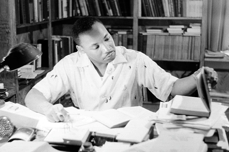 A black man sits at desk filled with papers and books.