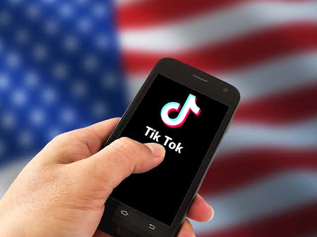 A person holds a smartphone with the TikTok logo on the screen, in front of a U.S. flag.