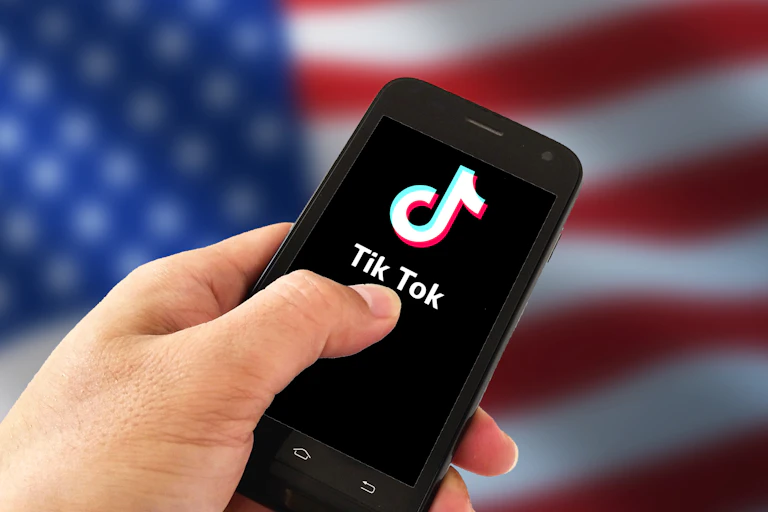 A person holds a smartphone with the TikTok logo on the screen, in front of a U.S. flag.