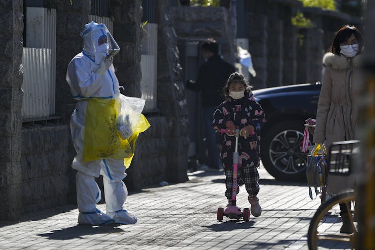 A child wearing a face mask and riding on a scooter passes by a worker in protective suit