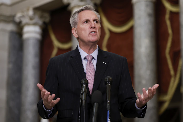 With statues in the background, a gray-haired man stands with his arms bent before him, palms face-up as he speaks.