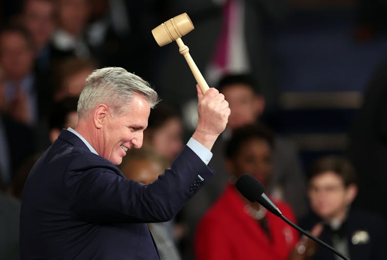 A gray-haired, smiling man in a suit lowers a gavel as people standing on the floor of the House of Representatives watch.