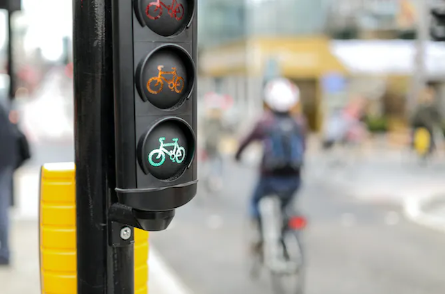 Lampu lalu lintas dengan sepeda hijau disorot di latar depan, dan seorang pengendara sepeda tidak fokus di latar belakang.