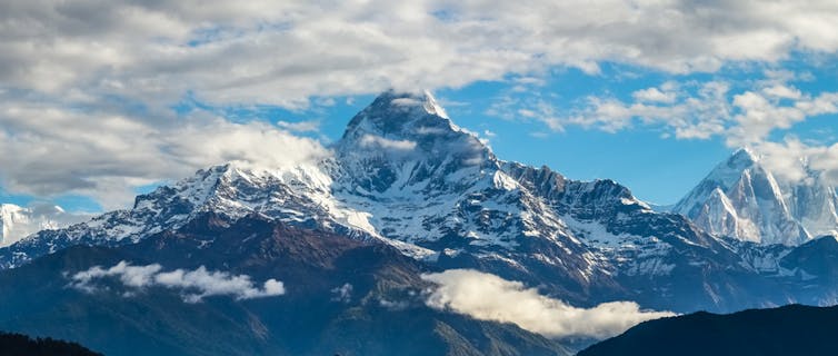 A picturesque landscape of snow capped jagged mountains seen from a distance, with a blue sky dappled with clouds