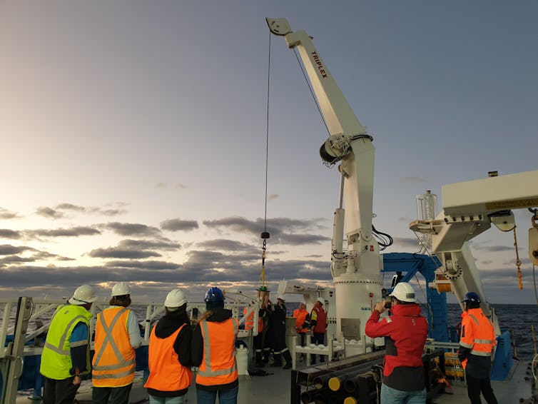 People on a ship in high-vis jackets watch a cable lower a scientific instrument into the sea
