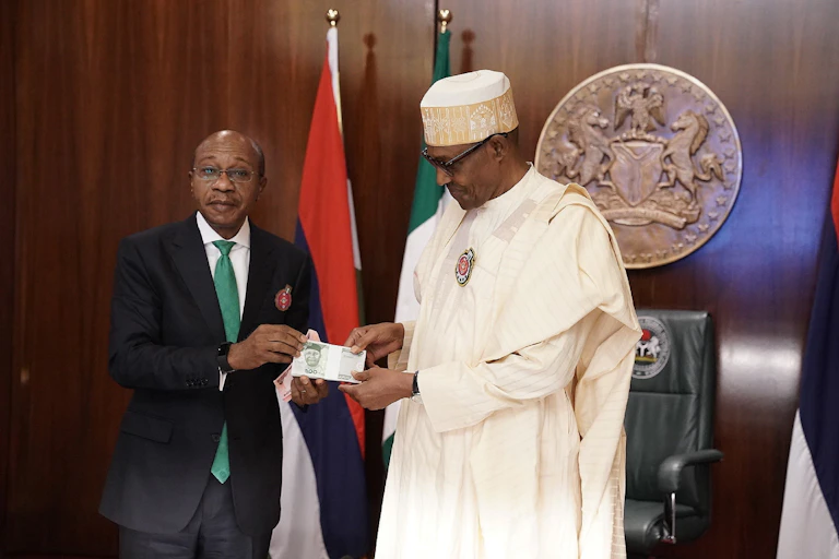 A man in a suit and a man in a robe and cap stand next to each other, holding a banknote, with a flag and seal behind them