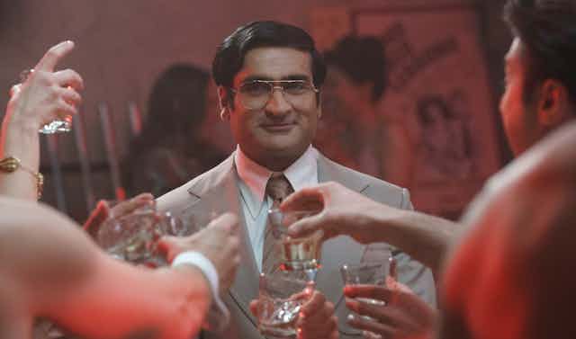 Kumail Nanjiani wears his hair in a side part, large glasses and a grey suit. Before him people are holding large shot glasses.