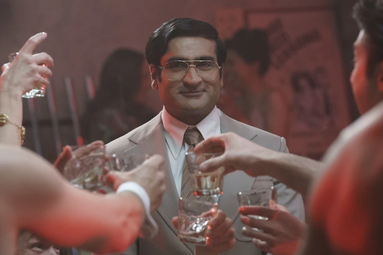 Kumail Nanjiani wears his hair in a side part, large glasses and a grey suit. Before him people are holding large shot glasses.