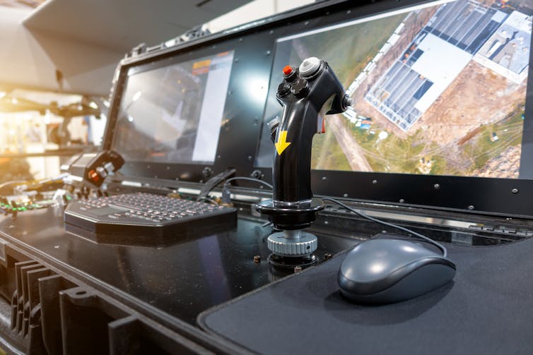 A joystick and control panel in front of a screen where a drone operation is taking place.