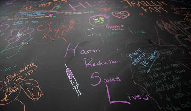 A wall with notes written in multi-coloured chalk. The one in the middle reads: Harm reduction saves lives. 