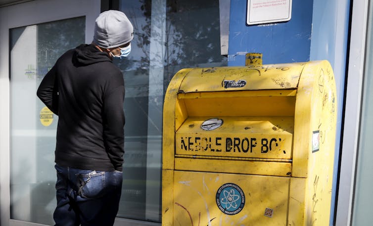 A man wearing a black jumper standing outside a building next to a large yellow bin labelled: needle drop box.