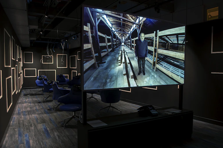 A large screen shows a man standing amid empty bunk beds in an old barracks.