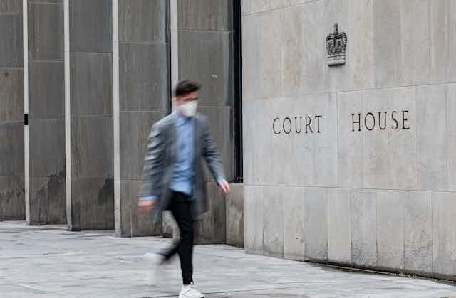 A man wearing a mask is blurred as he walks past a stone wall etched with the words Court House.