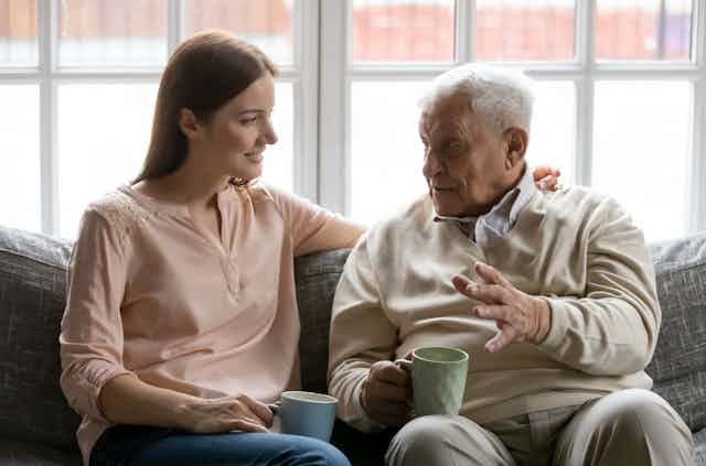 An elderly man talks to a young woman. They sit on a couch while carrying coffee mugs. 