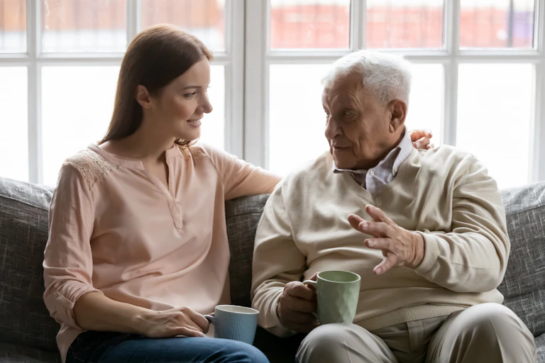 An elderly man talks to a young woman. They sit on a couch while carrying coffee mugs.