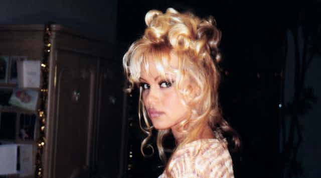 A young Pamela Anderson looks to camera wearing dark kohl eyeliner and her signature blonde hair piled high on her head. 
