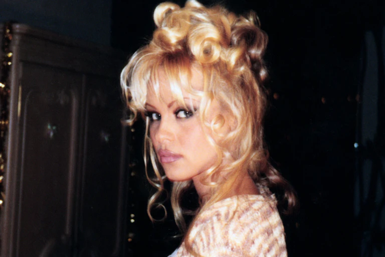 A young Pamela Anderson looks to camera wearing dark kohl eyeliner and her signature blonde hair piled high on her head.