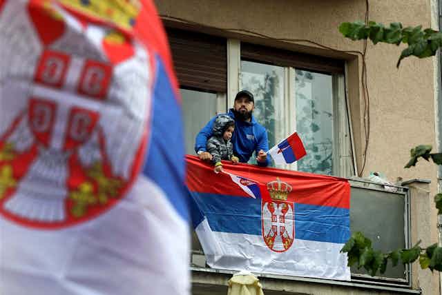 A man and a child stand on a balcony dressed in Serbian colours and waving Serbian flags.