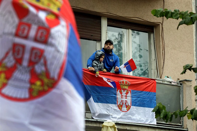 A man and a child stand on a balcony dressed in Serbian colours and waving Serbian flags.