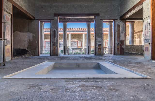 The atrium of the House of the Vettii, Pompeiii, showing a sunken pool, wide open column flanked doorways and a blue sky outside. 