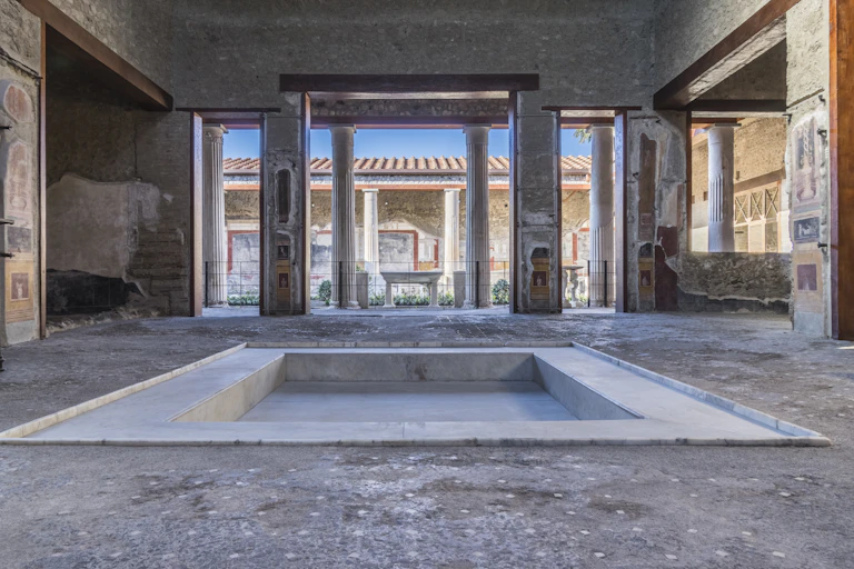 The atrium of the House of the Vettii, Pompeiii, showing a sunken pool, wide open column flanked doorways and a blue sky outside.