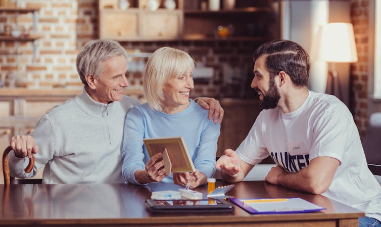 An elderly couple and a young man sit around a table chatting.