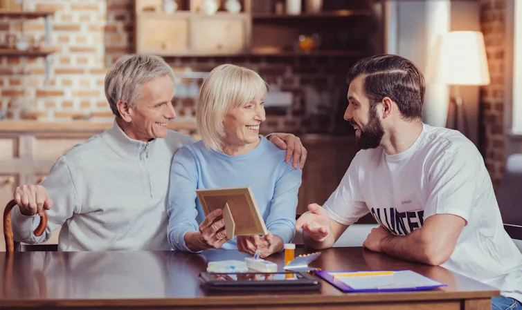 An elderly couple and a young man sit around a table chatting.
