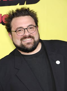 Kevin Smith in an all black suit wearing round glasses.