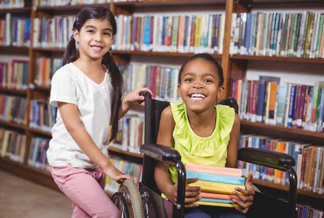 Two smiling girls in a school library. One is sitting in a wheelchair and the other is standing next to her.