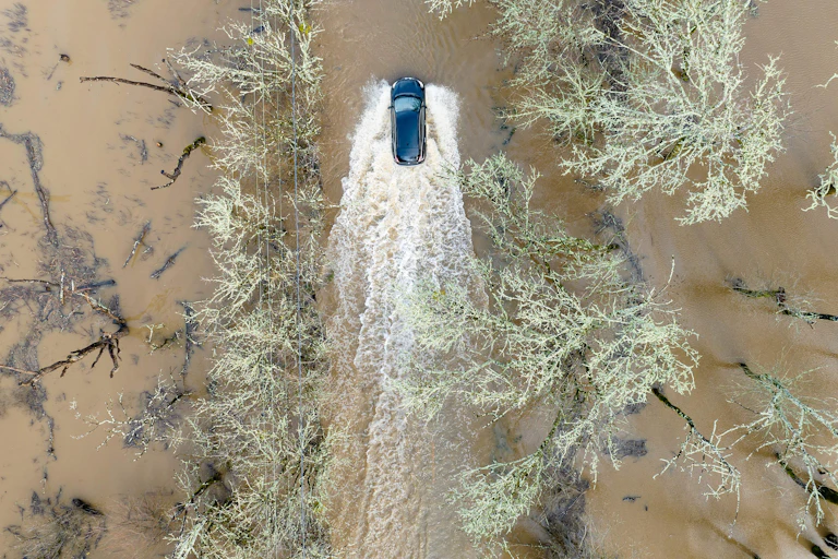 An SUV drives through a flooded road with muddy water on both sides and not much more than the trees and power lines to indicate where the road is.