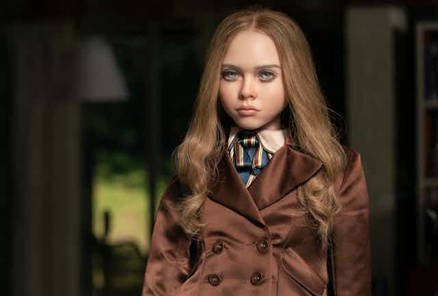M3gan the doll has long well placed hair and wears a trench coat.