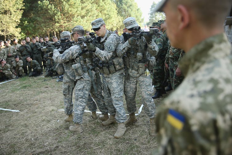 Soldiers in green camouflage appear to point guns in different directions, while other soldiers in dark green stand behind them and look on.