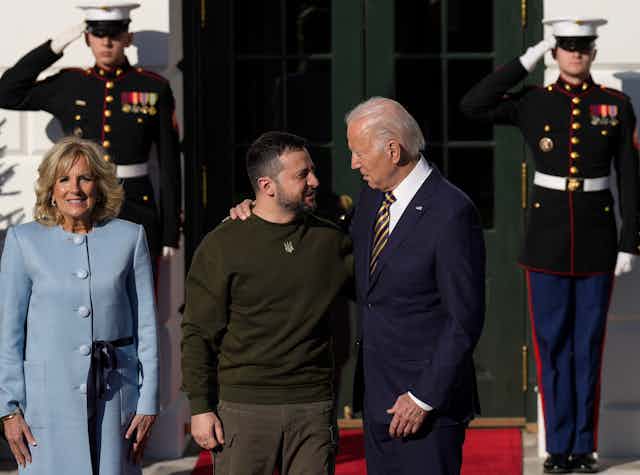 Two men face towards each other with their arms around each other. One is older and has on a blue suit. The other has short brown hair and green camo shirt. They stand in front of a woman with. blue suit and two soldiers behind them saluting. 
